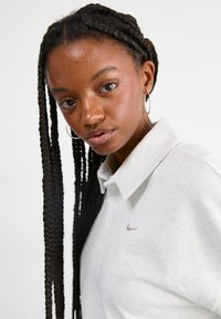 Šedé polo tričko s golierom a decentným logom Nike. Model má dlhé zapletené vlasy a nosí náušnice v tvare kruhu. Neutrálny pozadie.