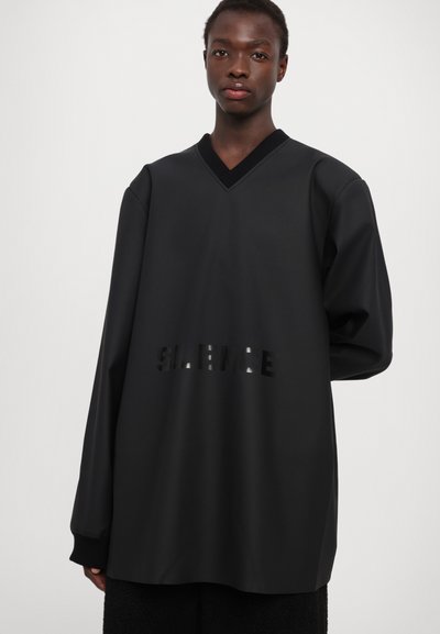 T-shirt noir oversize à manches longues avec un décolleté en V, arborant le mot "SILENCE" découpé de manière subtile sur le devant.