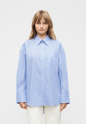 Ung kvinde med glat lyst hår iført en løs lyseblå skjorte med striber og hvide bukser mod en ensfarvet baggrund.