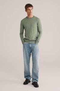 Grön långärmad tröja med en texturerad yta, kombinerad med ljusblå vida jeans och svarta skor. Modellen står med händerna sammanlänkade.