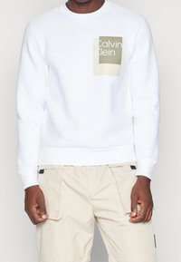 Sudadera blanca con escote redondeado y un parche de logo en verde y beige en el lado izquierdo del pecho. Combinada con pantalones cortos de carga en beige claro.