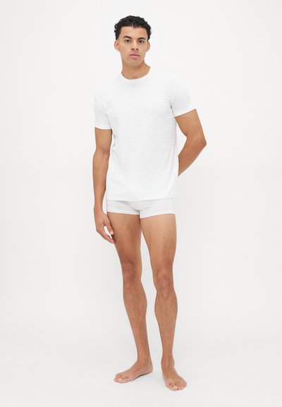Weißes, kurzärmeliges, strukturiertes T-Shirt kombiniert mit passenden weißen Unterhosen. Das Model steht barfuß und zeigt ein figurbetontes Design mit minimalen Details.