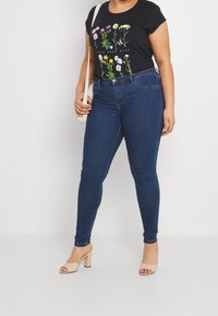 Svart t-shirt med blommig grafik; matchad med mörkblå skinny jeans. Modellen bär beige klacksandaler och har en ljus axelväska.