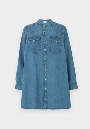 Femme portant une longue chemise en denim avec les manches retroussées, un jean bleu foncé à jambes larges et des talons noirs à bouts pointus, debout sur un fond blanc.