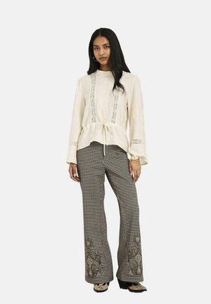 Mujer con el pelo largo y oscuro que lleva una blusa de encaje crema con lazo en la cintura y pantalones anchos estampados con bordado floral en los bajos.