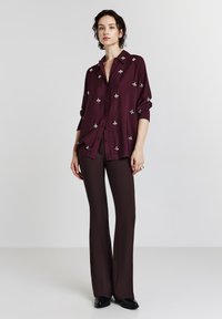 Donna che indossa una camicia a bottoni di un profondo viola con ricami bianchi a forma di ape e pantaloni a palazzo coordinati di un viola scuro, in piedi su uno sfondo neutro.