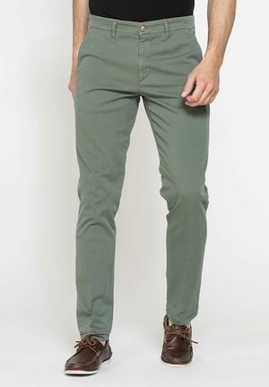 Pantaloni di cotone verde oliva con vestibilità slim, dotati di tasche frontali, chiusura con bottone e dettagli di cucitura sottili. Sono indossate scarpe marroni.
