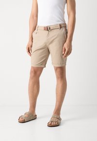 Shorts beige realizzati in tessuto leggero, con risvolti e una cintura intrecciata. Indossati con sandali grigi open-toe su uno sfondo neutro.