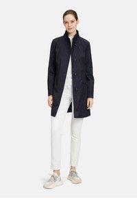 Manteau de trench en nylon imperméable de couleur marine avec un col montant, une fermeture à boutons, et deux poches avant, associé à un pantalon blanc et des baskets grises.