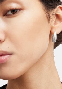 Boucle d'oreille en argent en forme de goutte d'eau avec une finition polie, comportant des détails gravés. Visible sur l'oreille d'un modèle sur un fond neutre.