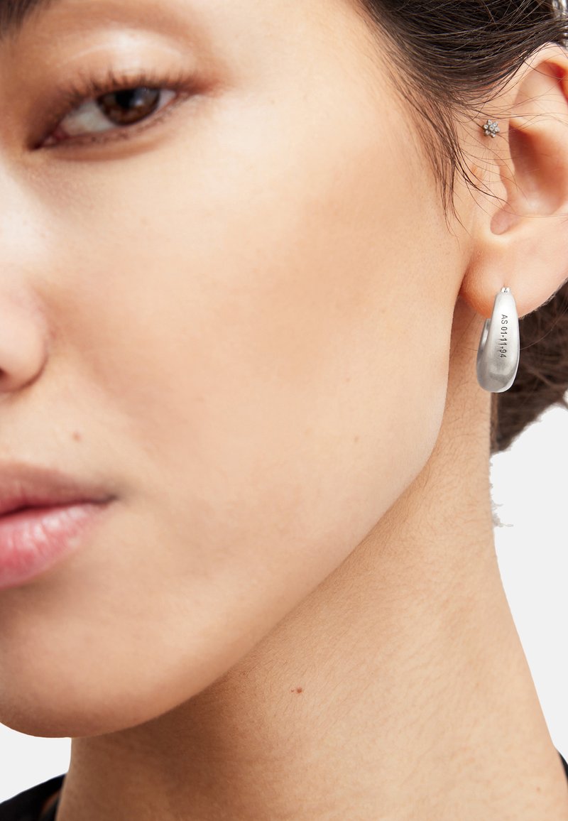 Boucle d'oreille en argent en forme de goutte d'eau avec une finition polie, comportant des détails gravés. Visible sur l'oreille d'un modèle sur un fond neutre.