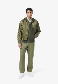 Giacca bomber verde sopra un hoodie verde, abbinata a pantaloni sportivi coordinati. Presenta una chiusura con zip, polsini a coste e sneaker bianche.