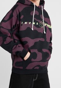Černá mikina s kapucí s růžovým a fialovým maskáčovým vzorem, obsahující klokaní kapsu, bílé šňůrky a výrazný zelený text loga na přední straně.