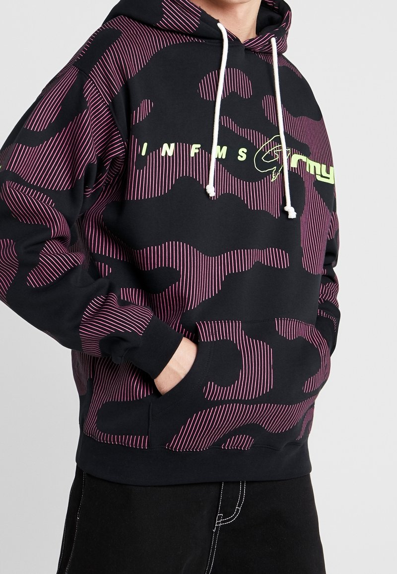 Černá mikina s kapucí s růžovým a fialovým maskáčovým vzorem, obsahující klokaní kapsu, bílé šňůrky a výrazný zelený text loga na přední straně.