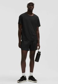 Musta lyhythihainen T-paita, mustat ruudulliset shortsit, mustat lenkkarit, valkoiset sukat ja valkoinen lippis, pitäen mustaa vesipulloa.