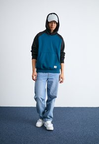 Felpa con cappuccio teal e nera con tasca frontale, abbinata a jeans azzurri e sneakers bianche. Presenta un cappellino e una vestibilità comoda.