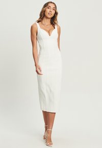 Witte midi-jurk met een aansluitend ontwerp, brede banden, een diepe halslijn en verticale naden. Vergezeld van sandalen met banden.