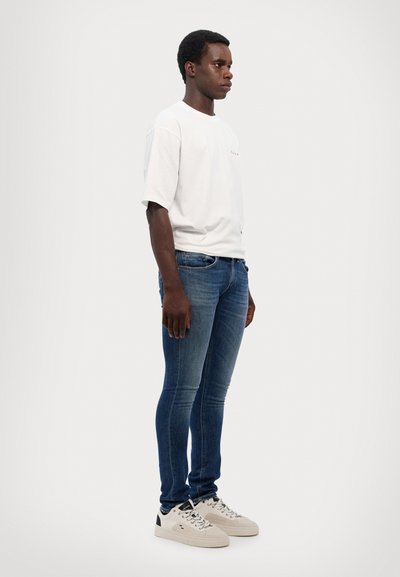 Outfit casual da uomo composto da una T-shirt bianca a maniche corte e jeans slim-fit blu scuro, abbinati a sneakers bianco sporco con dettagli neri.