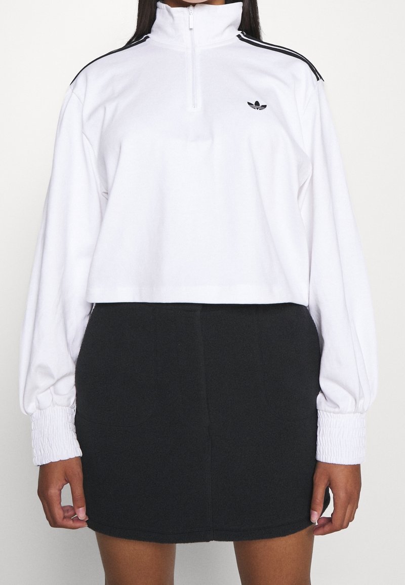 Osoba ubrana w biały krótki pulower z czarnymi paskami na ramionach oraz czarną spódnicę z wysokim stanem na jednolitym tle.