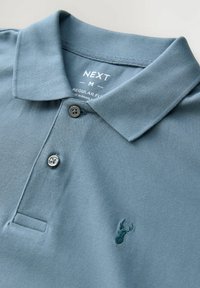 Camisa polo de tela de algodón en azul claro, con cuello, tapeta de dos botones y un pequeño logotipo de ciervo bordado en el pecho.