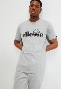 Camiseta de algodón gris con un logo negro de "ellesse". Manga corta, cuello redondo y corte relajado. Se lleva con pantalones grises a juego.