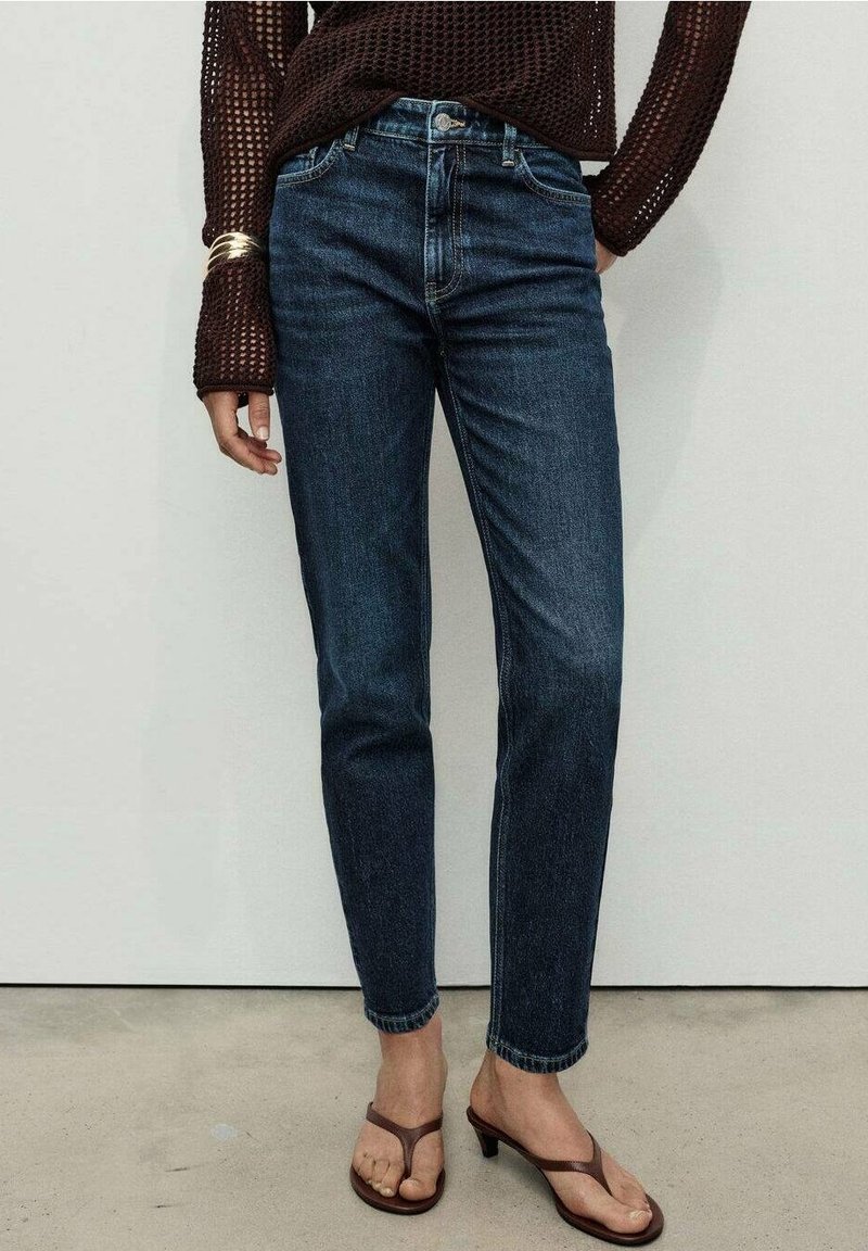 Personne portant un jean taille haute bleu foncé, un haut en mesh marron à manches longues, des bracelets dorés et des sandales à talons marron, debout sur un sol en béton.