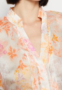 Col et encolure d'une blouse légère avec des motifs floraux oranges, roses et jaunes sur un tissu transparent.