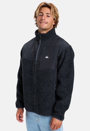 Giacca in pile di colore blu navy scuro, chiusura frontale con zip, colletto alto, pannello in tessuto nero con tasca, superficie testurizzata e polsini elasticizzati.