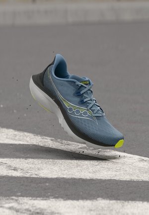 Chaussure de course bleue et noire avec semelle blanche et accents jaunes, flottant au-dessus d'une rue avec une ligne de passage piéton blanche.