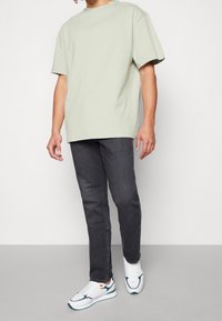 Persona con camiseta oversize verde claro, vaqueros ajustados negros y zapatillas blancas con detalles verdes y naranjas de pie sobre suelo gris.