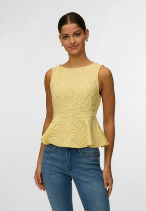 Femme portant un haut à basque sans manches jaune à fleurs, associé à un jean bleu, debout devant un fond clair uni.