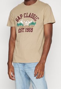 Béžové bavlněné tričko s grafikou hor a borovic, s nápisem "GAP CLASSIC EST. 1969" v vínově červeném písmu. Krátké rukávy, kulatý výstřih.