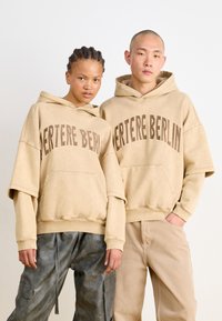 Felpe oversize beige con la scritta "BERLITE BERLIN" stampata in marrone scuro. Tessuto morbido, tasche a kangaroo, indossate con pantaloni cargo grigi e pantaloni beige chiaro.