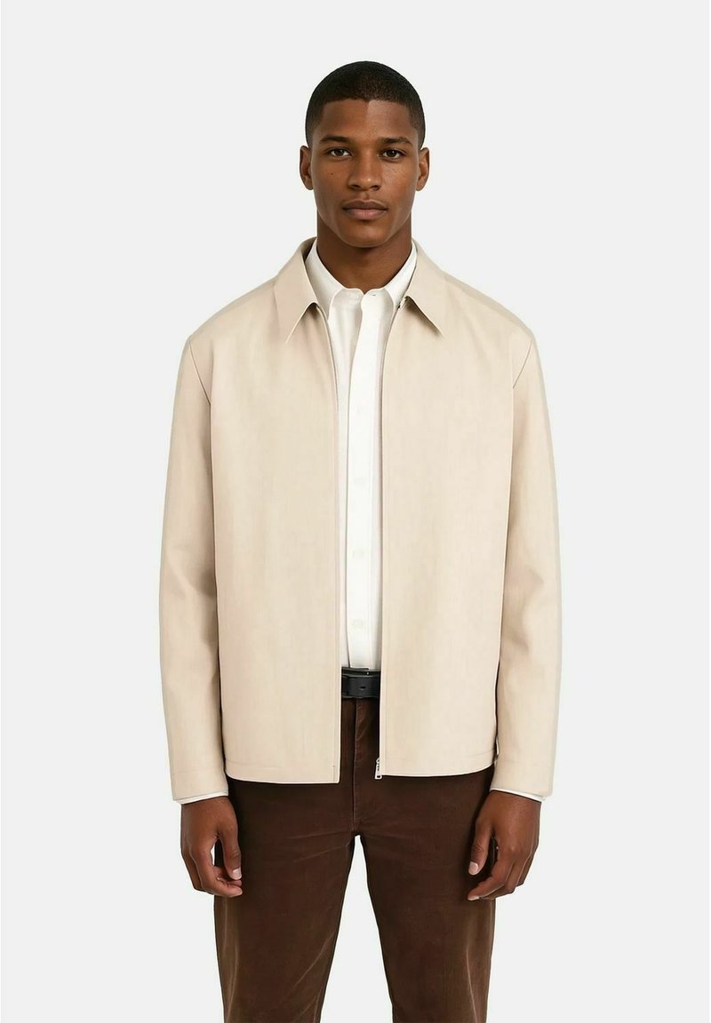 Beige zip-up jacka med en slät textur och spetsig krage, tillsammans med en vit skjorta med knappar och bruna byxor.