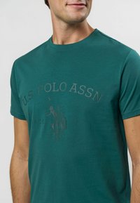 Zila kokvilnas T-kreklu ar smalku reljefu logo, kurā attēlots zirgs un jātnieks. Īsas piedurknes, apaļa kakla izgriezums un gluda tekstūra.