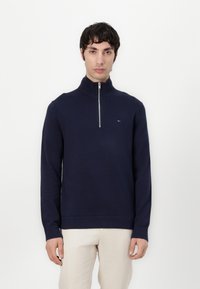 Marineblauwe gebreide zip-up trui met een hoge kraag en geribbelde manchetten. Beschikt over een klein logo op de borst. Zachte textuur.