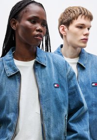 Dvaja jedinci majú na sebe rifľové bundy s svetlým praním, viditeľným prešívaním a prednými zipsami. Každá bunda má na hrudi malú nášivku s logom.