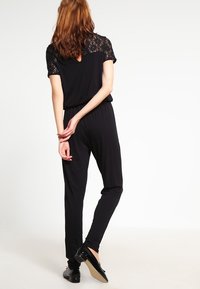 Černý overal s krajkovými krátkými rukávy, elastickým páskem a zúženými nohavicemi. Hladká textilie s jemným leskem. Zobrazeno ze zadní strany.