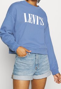 Sudadera azul con el logo blanco de "LEVI'S", diseño corto, cuello redondo y manga larga, combinada con pantalones cortos de mezclilla claros y con dobladillo.
