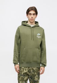 Oliivroheline kapuutsiga sviiter, mis on valmistatud puuvilla segust. Sellel on känguru tasku, reguleeritav kapuuts ja valge trükitud logo rinnal.