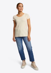 Beige T-Shirt mit rundem Ausschnitt und kurzen Ärmeln, kombiniert mit blauen, gerade geschnittenen Jeans und hellen Sneakers.