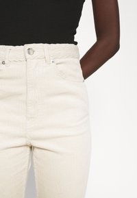 Jeans de veludo cotelê bege com cintura alta, apresentando dois bolsos frontais, fecho de botão e listas verticais texturizadas.
