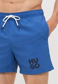 Modré plavky s elastickým pásom a bielou šnúrkou, čiernym potlačeným logom, hladkou štruktúrou a strihom nad kolená.