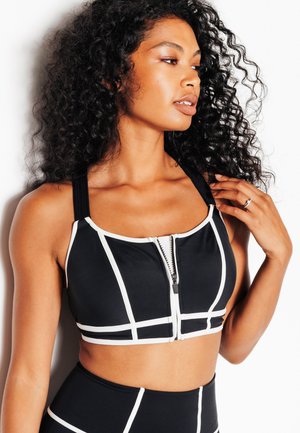 Donna con capelli ricci che indossa un reggiseno sportivo nero con bordi bianchi e una zip frontale, toccandosi i capelli e guardando di lato su uno sfondo bianco.