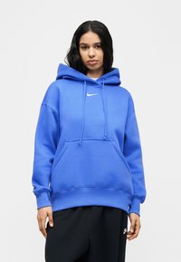 Γυναίκα με μακριά σκούρα μαλλιά, που φοράει μπλε φούτερ με κουκούλα Nike και μαύρο παντελόνι Nike, στέκεται μπροστά σε ένα απλό ανοιχτό φόντο.