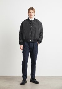 Fekete bomber dzseki patent gombokkal, fehér ing fölött viselve. Tengerészkék cargo nadrággal és fekete fűzős cipővel párosítva. Simább textúra.