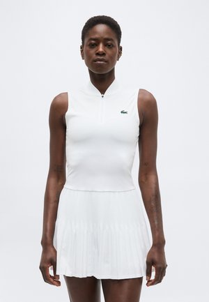 Naine kannab valget varrukateta lukuga särkriiet rohelist krokodillilogot ja valget volditud seelikut tavalise taustaga.