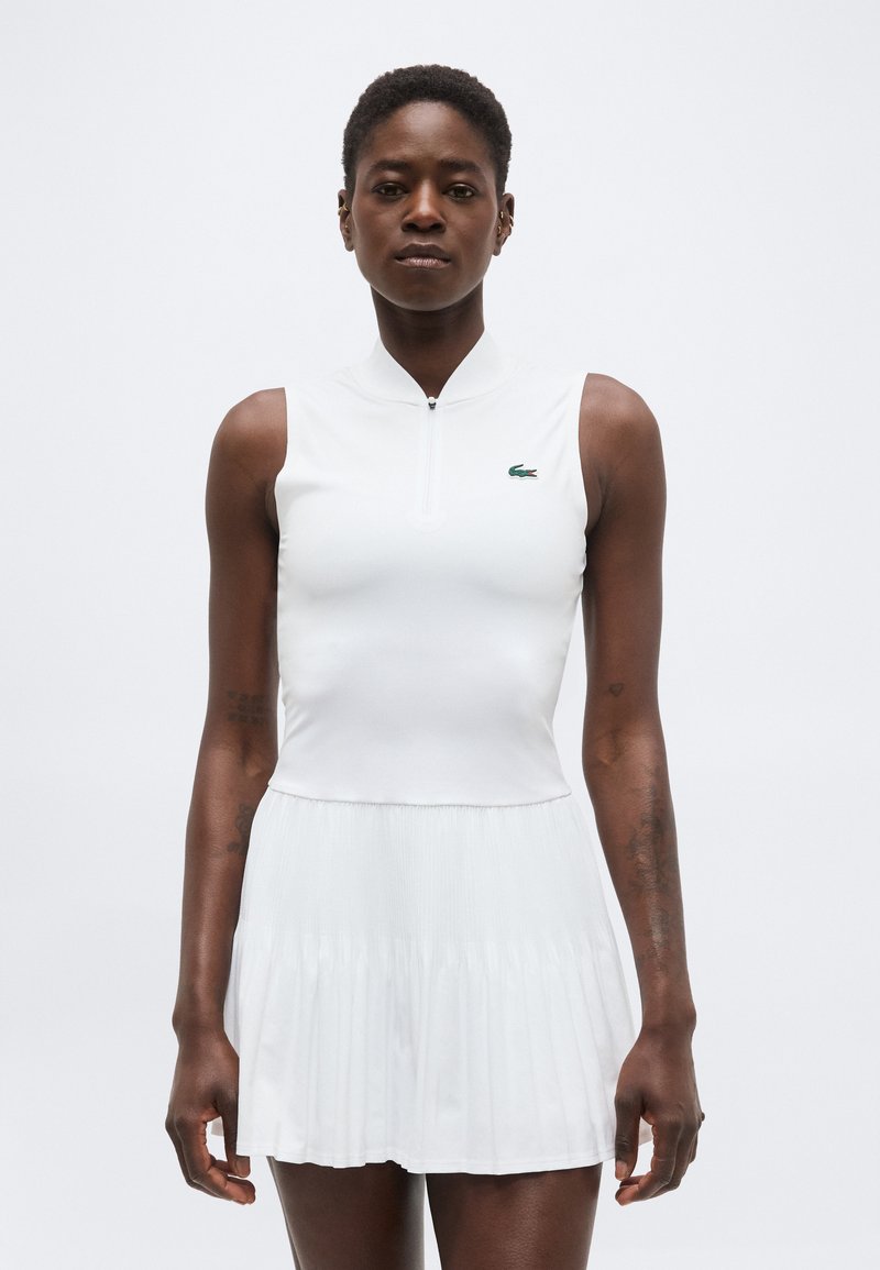 Γυναίκα φορώντας λευκό αμάνικο τοπ με φερμουάρ και πράσινο λογότυπο κροκόδειλου και λευκή πλισέ φούστα σε απλό φόντο.