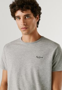 Ung mand med krøllet brunt hår iført en lysegrå Pepe Jeans London t-shirt, der kigger let til siden mod en ensfarvet baggrund.