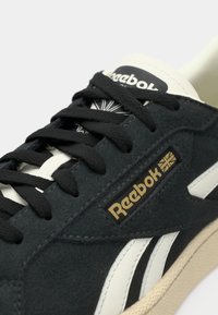 Crne suede tenisice s crnim vezicama, bijelim detaljima i zlatnim logotipom. Imaju teksturiranu površinu i nizak profil.